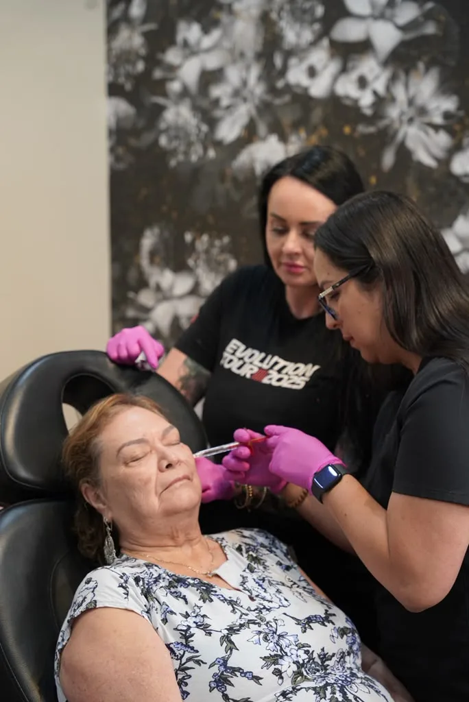 A trainee injecting a client's cheek, guided by the trainer at her shoulder