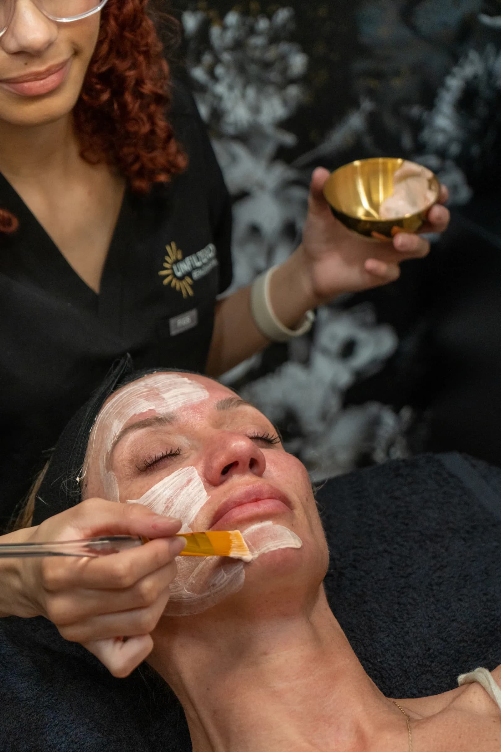A peel being brushed onto a client's face from a gold bowl
